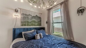 Une chambre à coucher avec un lit, une lampe fixée au mur, une fenêtre avec des rideaux et une plante suspendue.