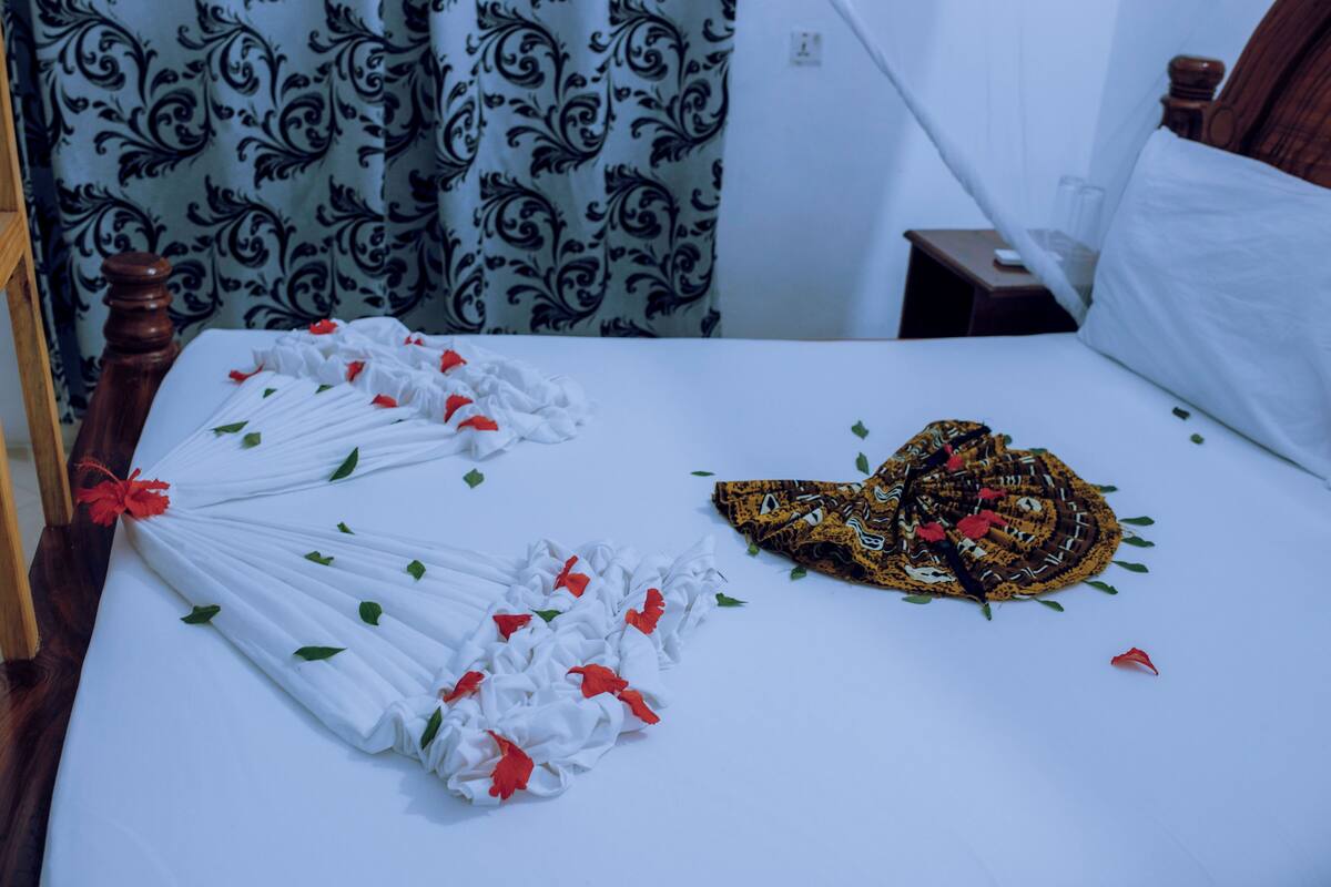Una cama bien tendida con cubrecama blanco, un cabecero decorativo y una cortina estampada al fondo.