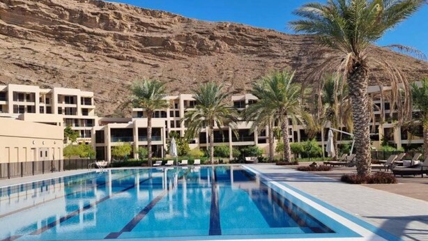 A swimming pool with clear blue water, surrounded by palm trees and modern buildings with balconies. -  Two Bedroom Apartment Muscat Bay (Bandar Jissah)