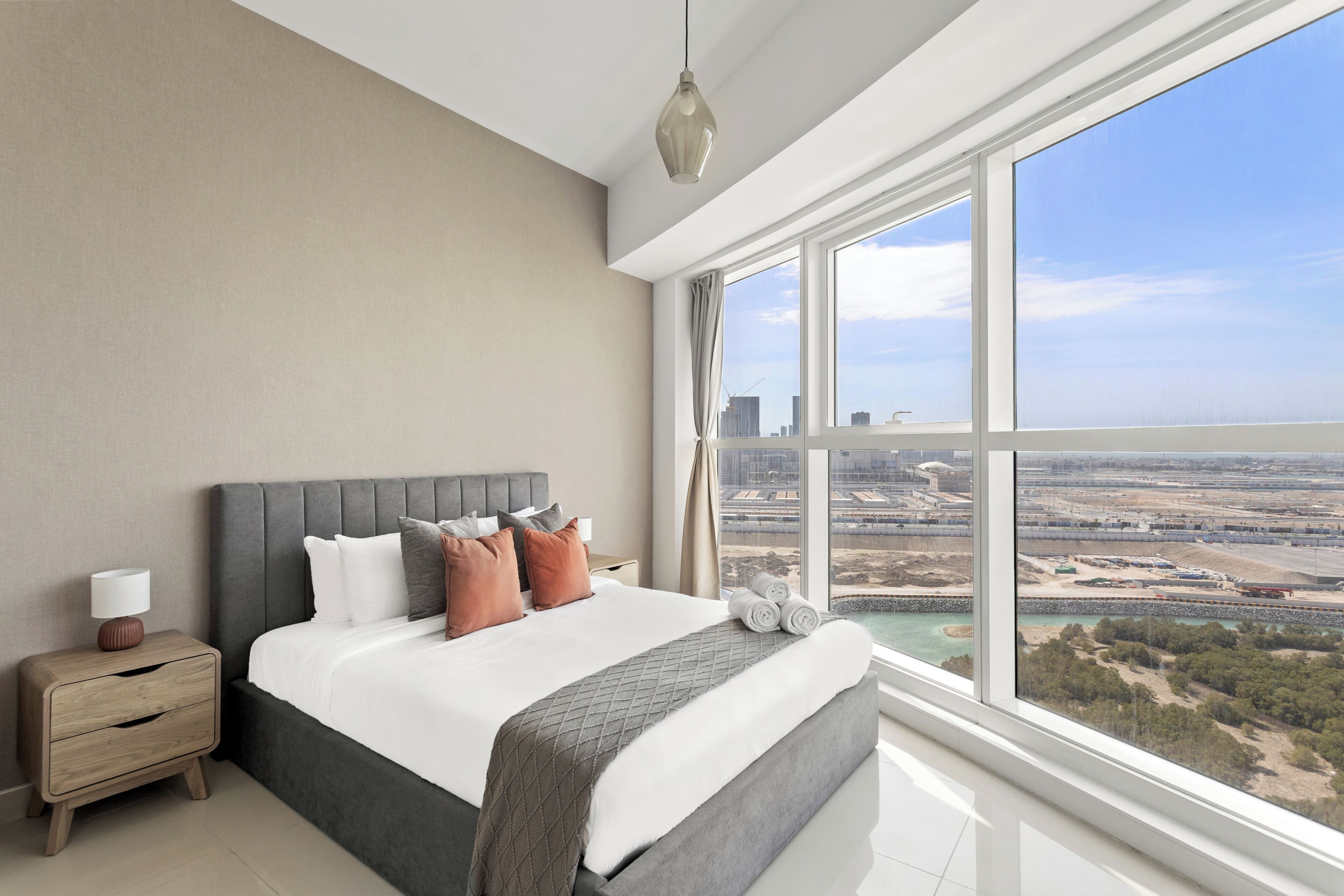 A modern bedroom with a large bed, a nightstand, and a view of a cityscape through large windows.