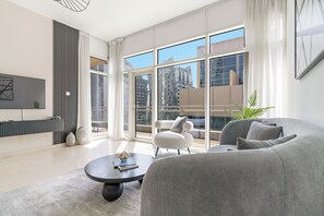 A modern living room with a grey sofa, a round coffee table, and a white armchair. Large windows offer a view of tall buildings. - Silkhaus Bay Square 13 (Dubai)