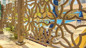 A decorative metal fence with a geometric pattern, a pool area with lounge chairs and umbrellas, and tall buildings in the background. - Silkhaus Amwaj 4 (Dubai)