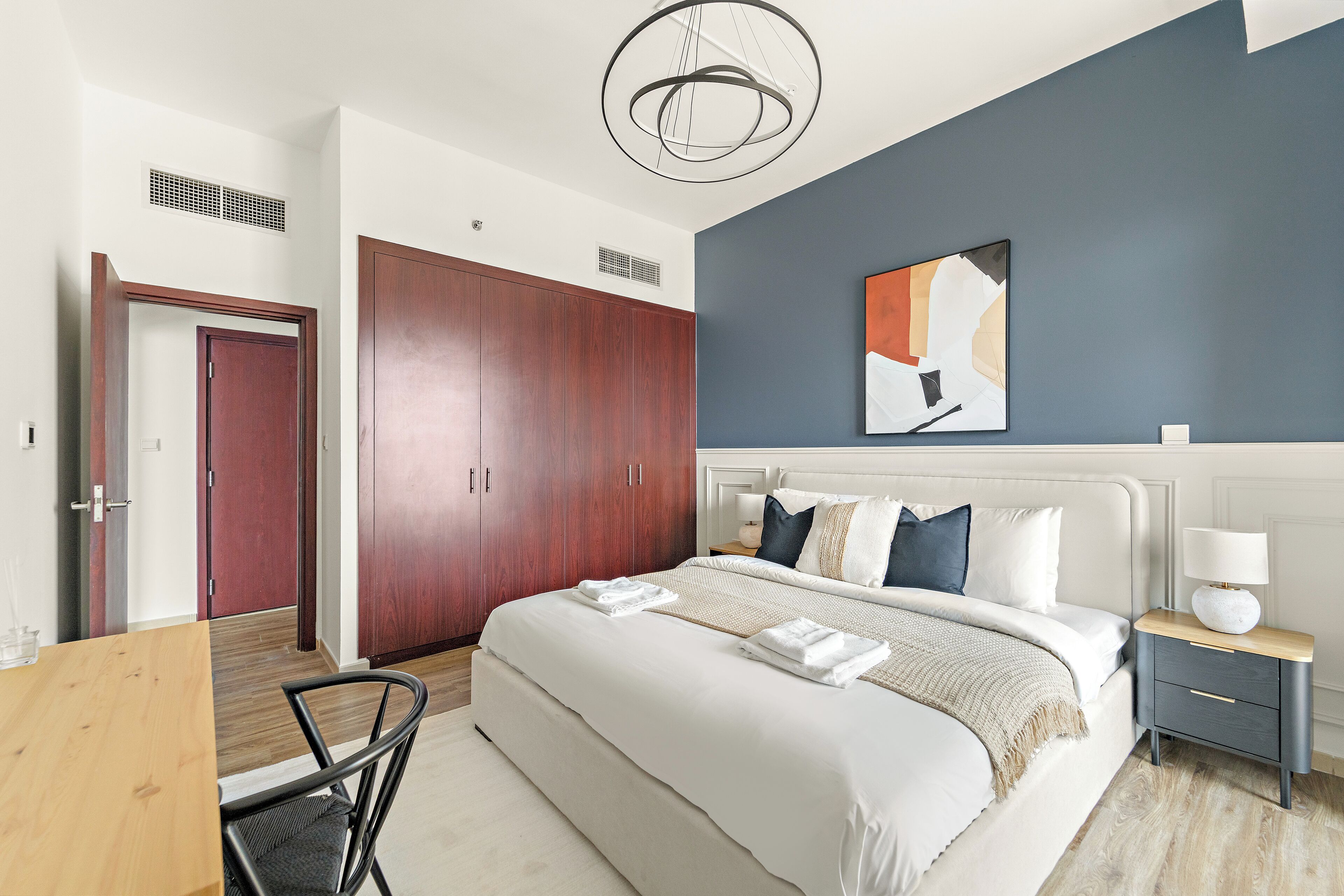 A modern bedroom with a large bed, a wooden wardrobe, a bedside table with a lamp, and a ceiling light fixture.