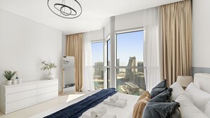 A modern hotel room with a large window, a white bed, a blue throw, and a white dresser with plants.