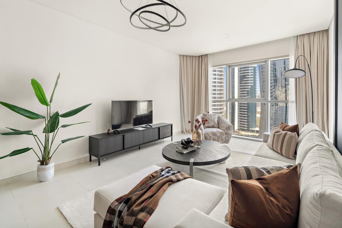 A modern living room with a sofa, coffee table, and a TV. There is a large window with curtains, a potted plant, and a city view.
