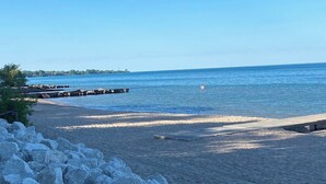 Ein Strand mit steiniger Uferböschung, einer Pier, die sich in ruhiges Wasser erstreckt, und einem wolkenlosen Himmel.