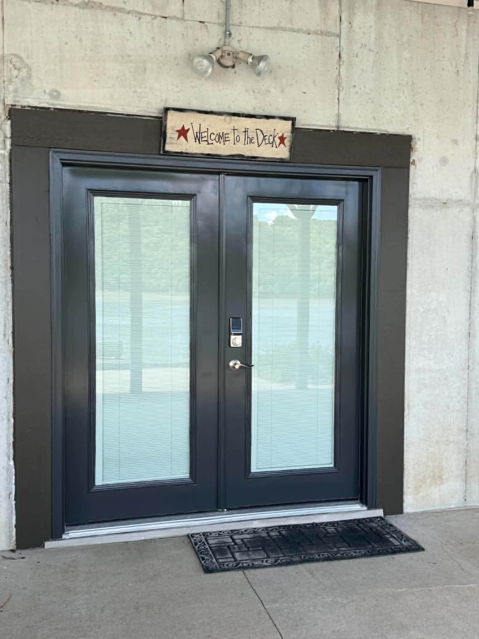 A black double door with a frosted glass panel, a 'Welcome to the Deck' sign above, and a doormat at the entrance.
