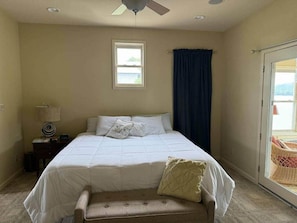 A bedroom with a bed, a nightstand, a lamp, a window, and a view of the outdoors.