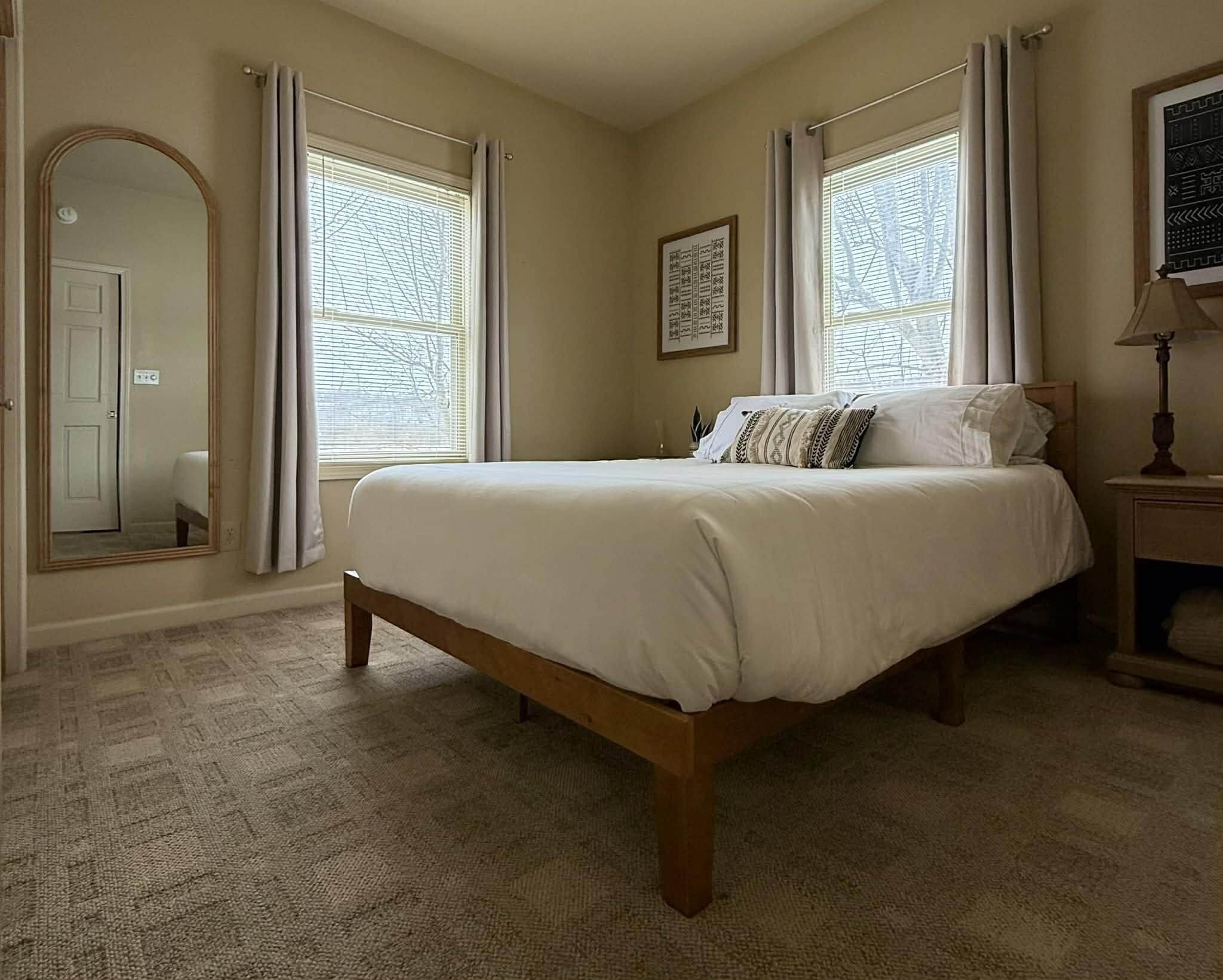 Quarto com cama grande, duas janelas com cortinas, mesa de cabeceira com abajur e um quadro na parede.