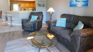 A living room with a sofa, armchairs, a glass coffee table, and a lamp. There is a kitchen in the background with wooden cabinets and a bar counter. - Gardens of Beachwalk 1/1 with Balcony (Fort Myers)