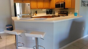 A kitchen with wooden cabinets, stainless steel appliances, a central island with bar stools, and a painting on the wall. - Gardens of Beachwalk 1/1 with Balcony (Fort Myers)