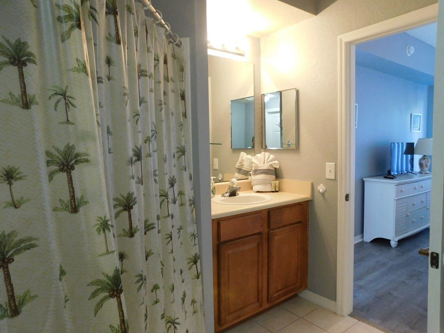 A bathroom with a shower curtain featuring palm tree patterns, a sink with a mirror above it, and a vanity with a lamp and decorative items.