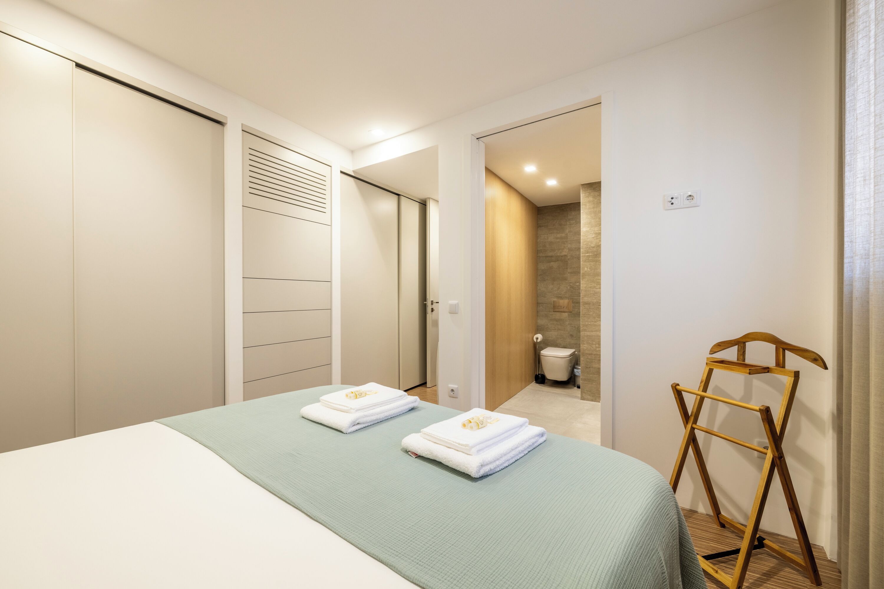 Une chambre d’hôtel avec un lit, un étendoir en bois et une salle de bain visible à travers une porte ouverte.