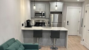 Eine moderne Küche mit grauen Schränken, einem Edelstahlkühlschrank und einer Mikrowelle. Ein Türkis Sofa und zwei Barhocker sind ebenfalls vorhanden.