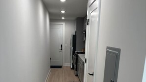 Un couloir étroit aux murs de couleur claire et au plancher en bois, qui mène à un espace cuisine équipé d’un réfrigérateur et d’un four à micro-ondes.