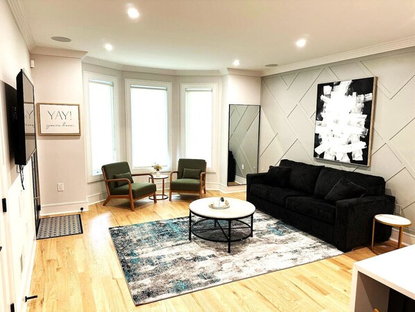 Un salon moderne avec un canapé noir, deux fauteuils verts, une table basse et un grand tableau abstrait accroché au mur.