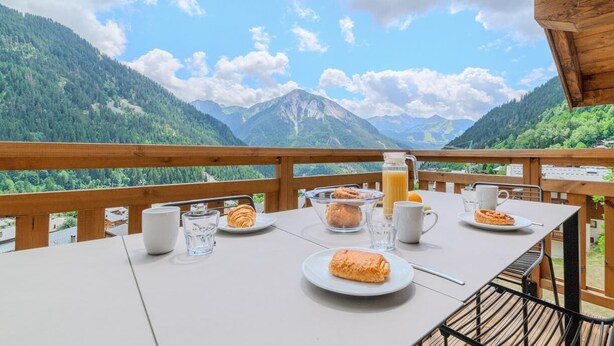 Tavola imbandita per la colazione su un balcone con vista sulle montagne, con un bicchiere di succo, croissant e pasticcini.