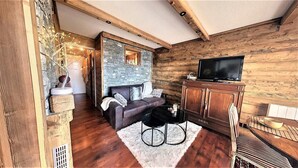 Una sala de estar acogedora con pared de piedra, vigas de madera, un sofá de cuero, una mesa de centro negra y un televisor sobre un mueble de madera.