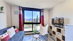 A living room with a sofa, a white chair, a TV, and a view of mountains through a large window.