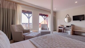 A hotel room with a bed, two armchairs, a desk with a chair, a TV, and a balcony with a view.
