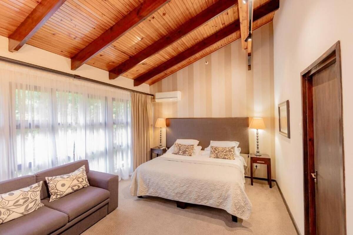 Un dormitorio con techo de madera, cama con almohadas estampadas, mesita de noche y ventana con cortinas.