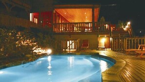 A swimming pool with a wooden deck, a covered outdoor seating area, and a building with a balcony.
