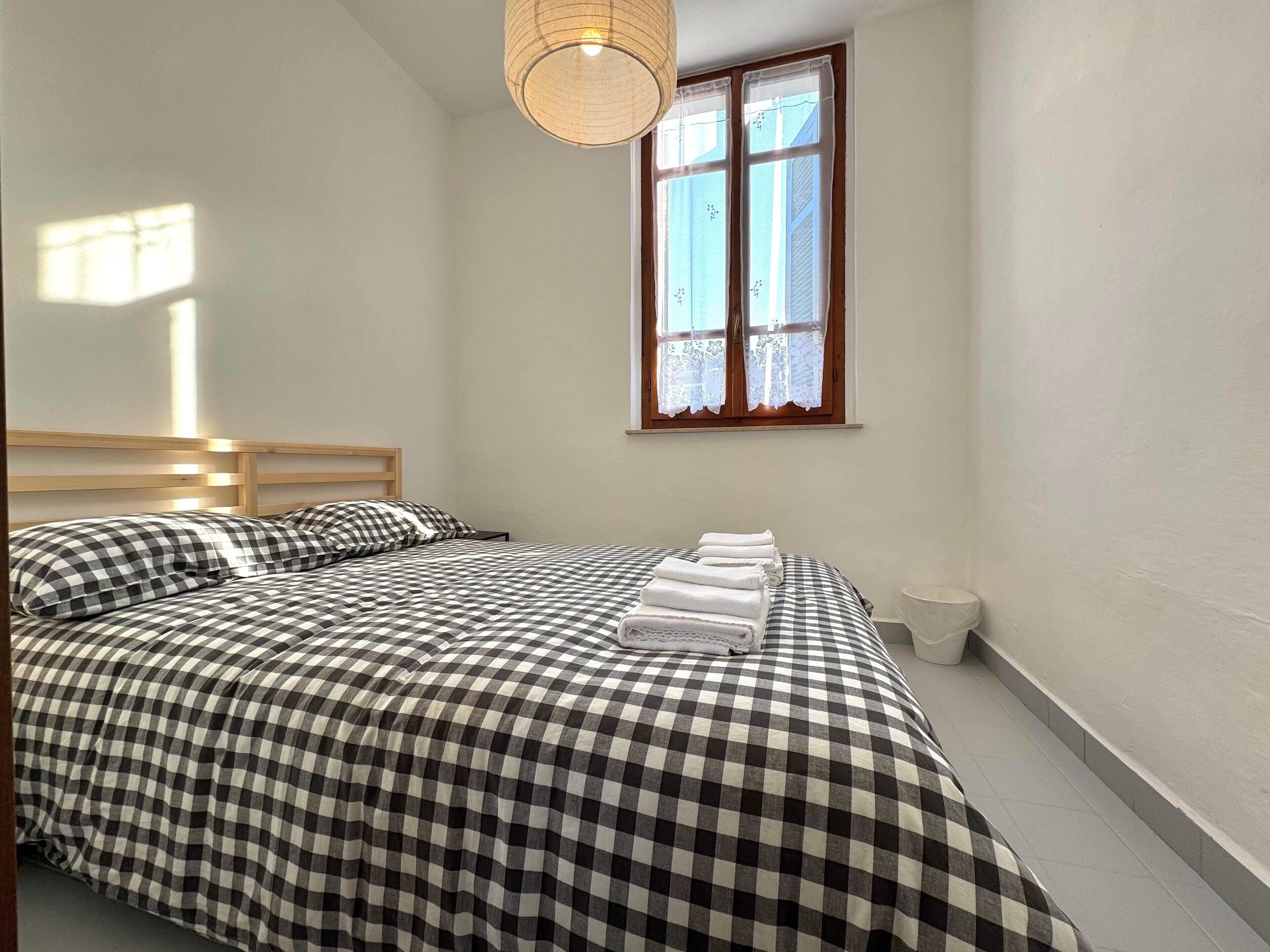 A bedroom with a checkered bedspread, a wooden headboard, a window with lace curtains, and a bedside lamp.