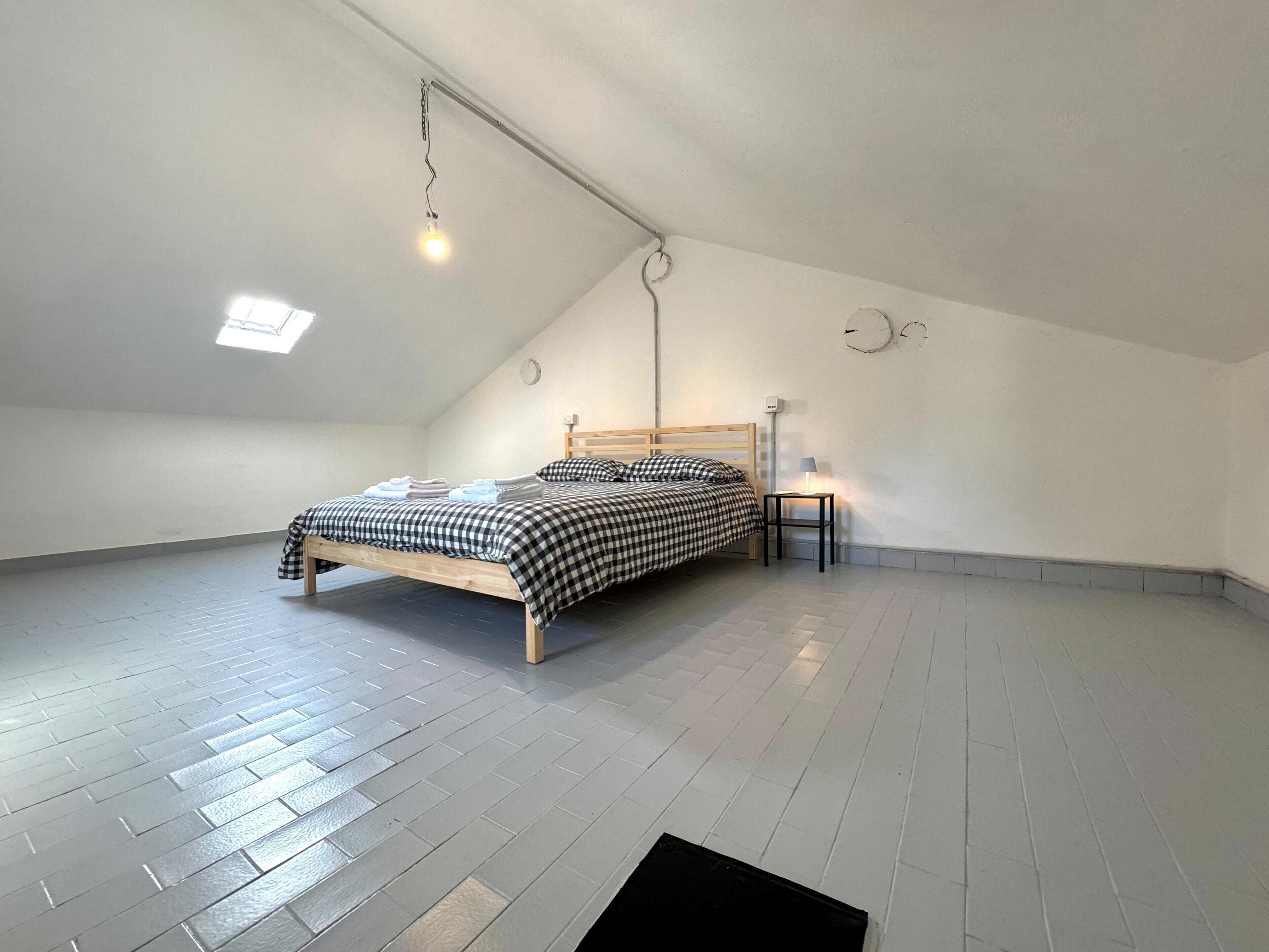 Une chambre spacieuse située dans les combles, avec un plafond en pente, un lit en bois, une petite table de chevet avec une lampe et un puits de lumière.