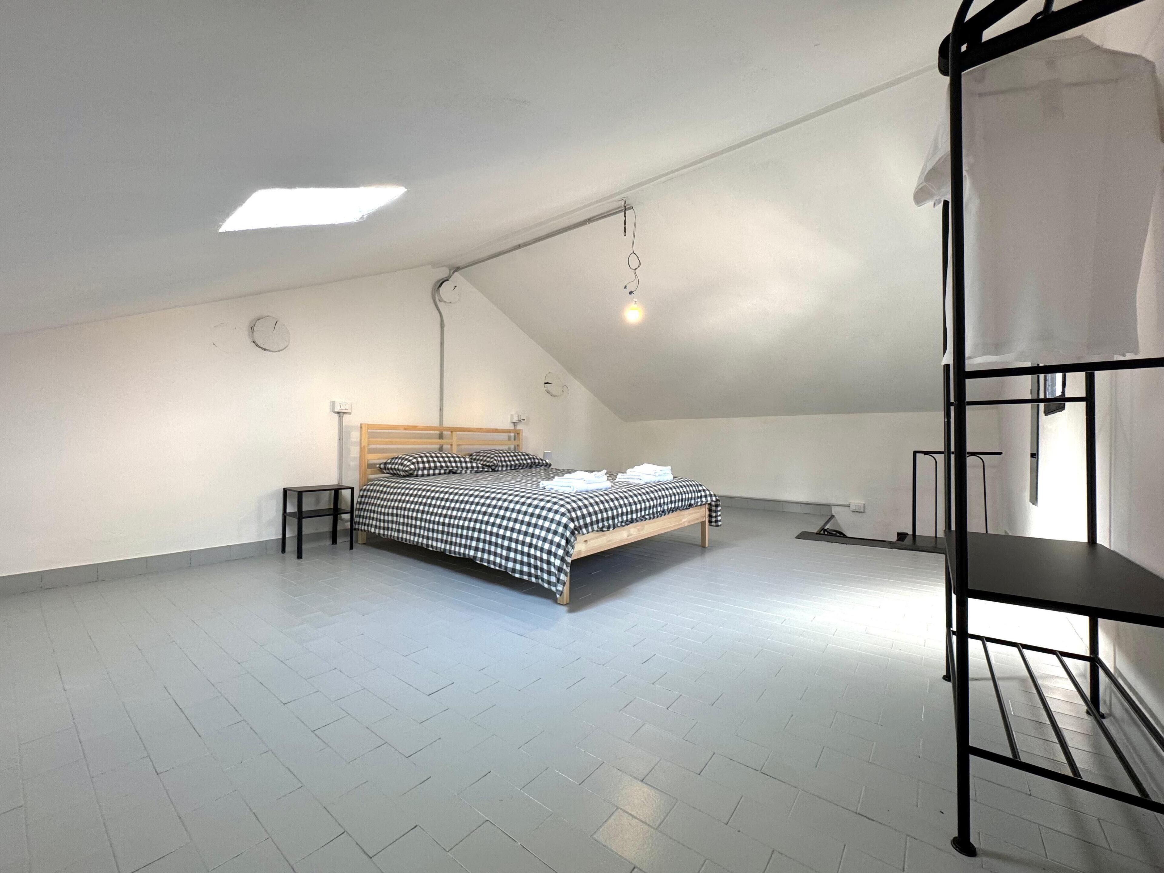 A spacious attic bedroom with a sloped ceiling, a bed with checkered bedding, a small bedside table, and a bunk bed on the right.