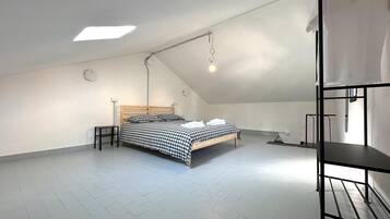 A spacious attic bedroom with a sloped ceiling, a bed with checkered bedding, a small bedside table, and a bunk bed on the right.