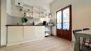A compact kitchen with white cabinets, a microwave, and a coffee maker. There is a dining table with chairs and a wooden door with a lace curtain. - Via Donati - O&R Home Crema (Crema)