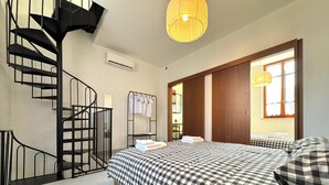 A bedroom with a checkered bedspread, a black spiral staircase, a hanging light fixture, and a window with a view. - Via Donati - O&R Home Crema (Crema)