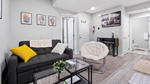 Un salon moderne avec un canapé noir, un fauteuil blanc et une table basse en verre. Des tableaux encadrés ornent le mur, et une porte donne accès à une autre pièce.