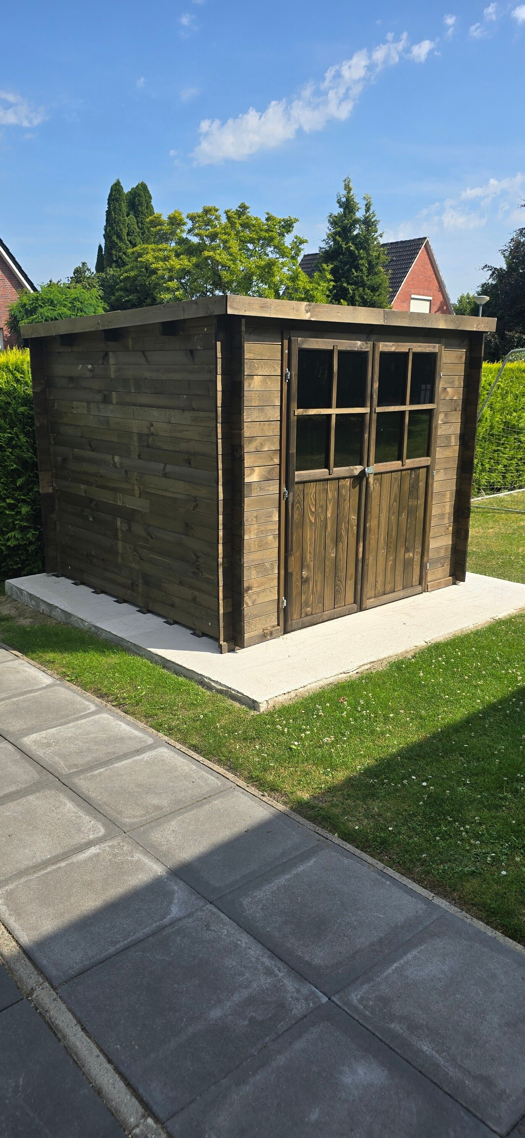 Un abri en bois doté d’une porte et de fenêtres, installé sur une dalle de béton et bordé d’une zone herbeuse.
