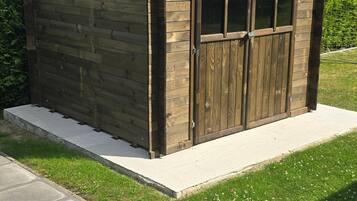 Un abri en bois doté d’une porte et de fenêtres, installé sur une dalle de béton et bordé d’une zone herbeuse.