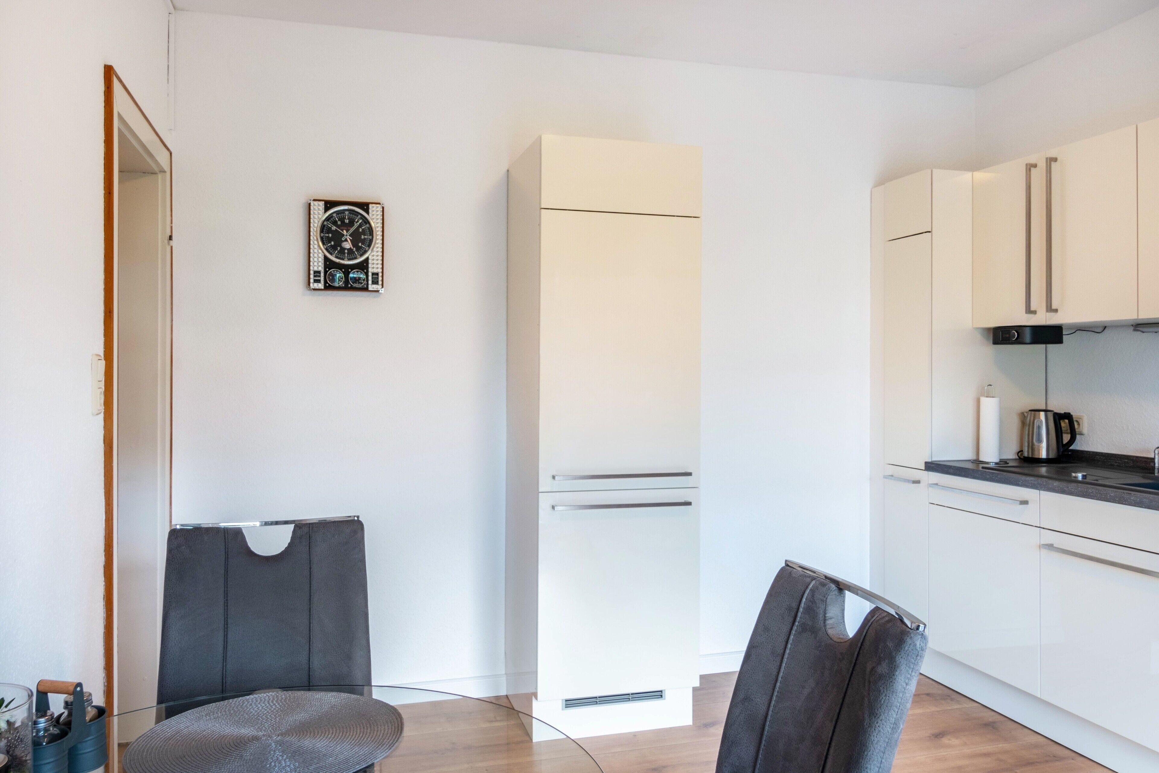 A modern kitchen with white cabinets, a refrigerator, a clock, and a dining area with a table and chairs.