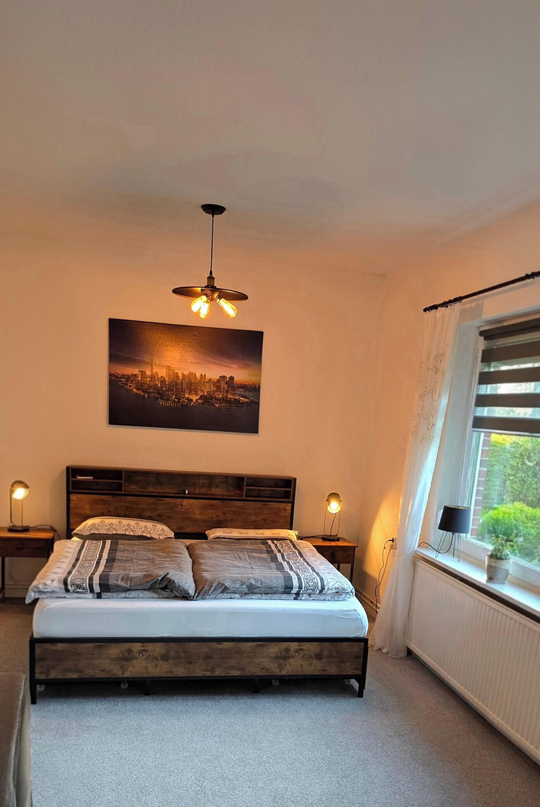 Un dormitorio con cama, un cuadro de paisaje urbano, una lámpara en el techo, una ventana con cortinas y un radiador.