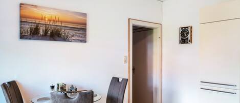 A room with a dining table, two chairs, a painting of a sunset, a clock, and a wardrobe.