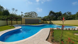 A swimming pool with a curved design, surrounded by a stone patio and bordered by a grassy area with red flowers. - Barmel Apartments (Coxen Hole)