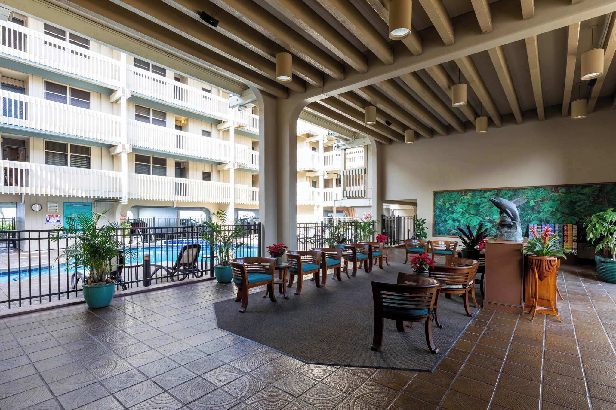 Un espacio al aire libre cubierto con sillas y mesas de madera, una piscina al fondo y vistas a un edificio de varios pisos con balcones.