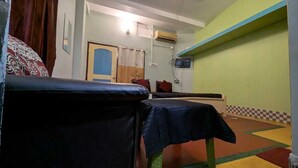 A room with a sofa, a small table with a tablecloth, a door with a curtain, and an air conditioning unit on the wall. - Baikuntha Dham Bhakta Niwas (Puri)