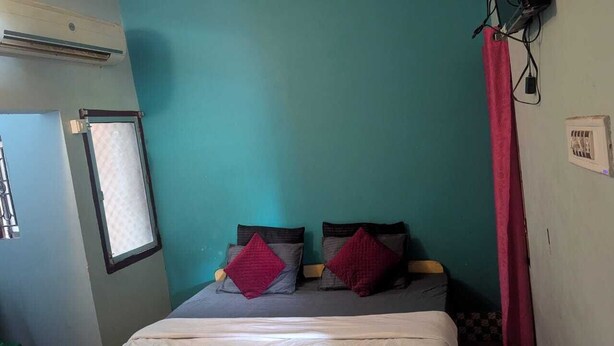 A room with teal walls, a bed with a white mattress and two pillows, a window with a blind, and a red curtain. - Baikuntha Dham Bhakta Niwas (Puri)