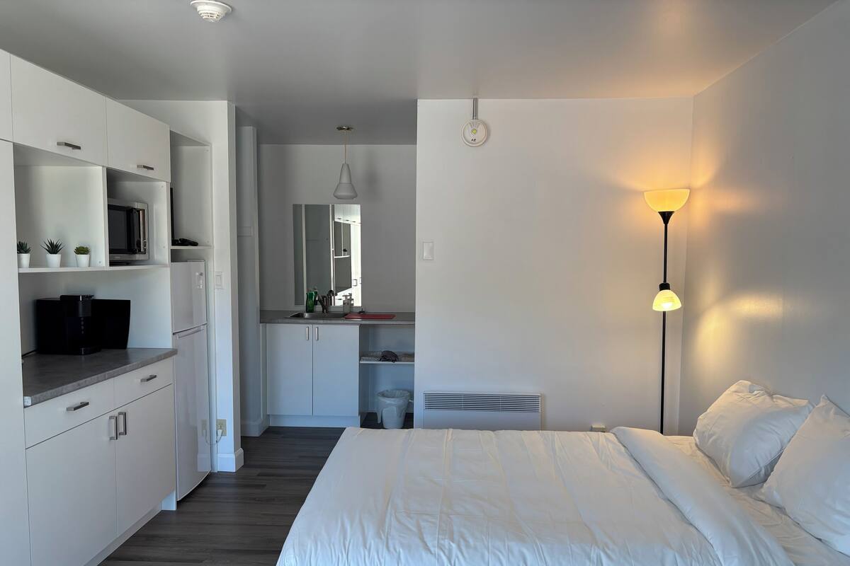 A modern bedroom with a large bed, a kitchenette with a microwave, and a bedside lamp.
