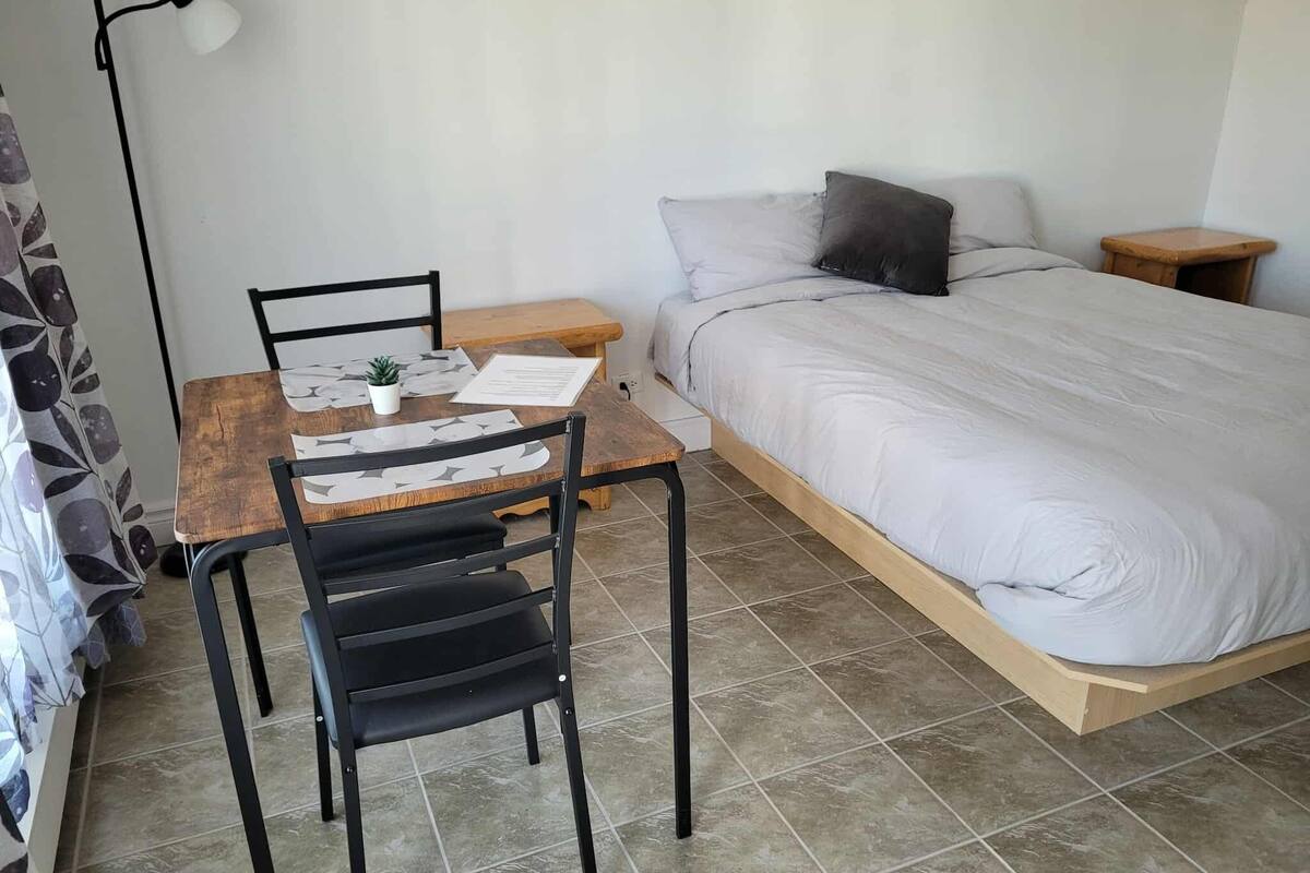 A bedroom with a bed, a small table with a chair, a bedside table, and a wall-mounted light.