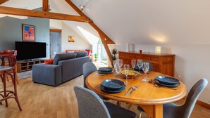 Un coin repas avec une table en bois dressée pour quatre personnes, entourée de chaises grises. On aperçoit un téléviseur à l’arrière-plan, et la pièce est dotée d’un plafond en pente avec des poutres apparentes.