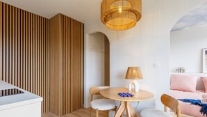 A modern living space with a round wooden table, two chairs, a lamp, and a bed with a pink blanket.