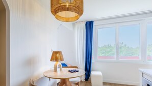A modern room with a round wooden table, a lamp, a chair, a small white ottoman, and a view of greenery through a window with blue curtains.