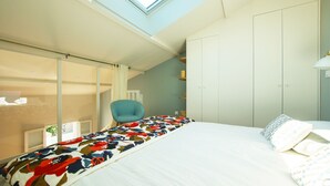 A bedroom with a skylight, a bed with a colorful blanket, a blue chair, and a wardrobe.