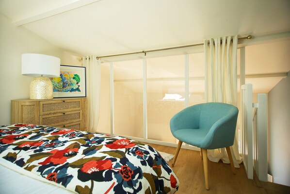 Une chambre à coucher comprenant un lit, une chaise, une commode et une grande fenêtre avec des rideaux.