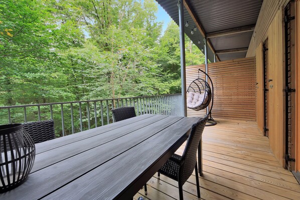 Una terraza de madera con una mesa y sillas, un sillón colgante y vistas a un área boscosa.